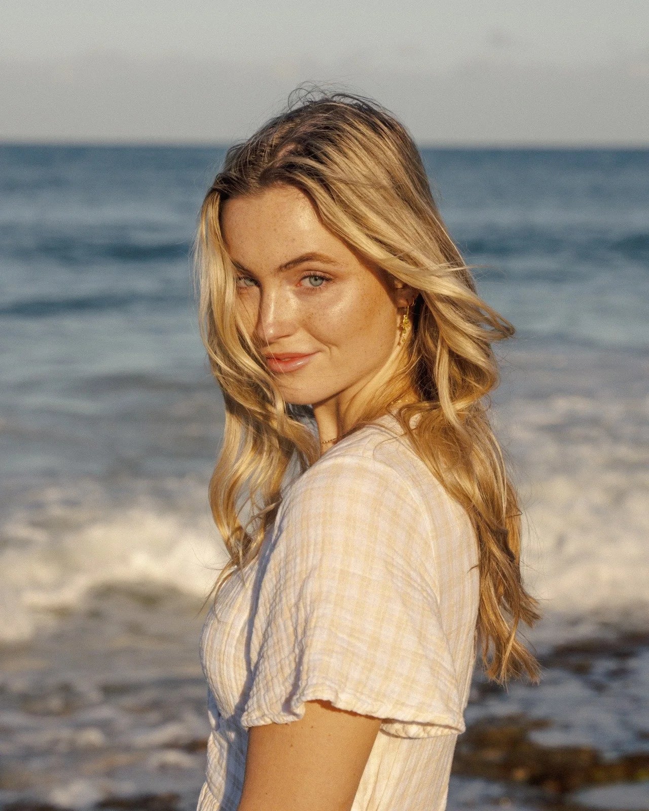Blonde woman smirking at the camera with the ocean behind her, evoking Oshun’s connection to nature and luxury skincare lifestyle.