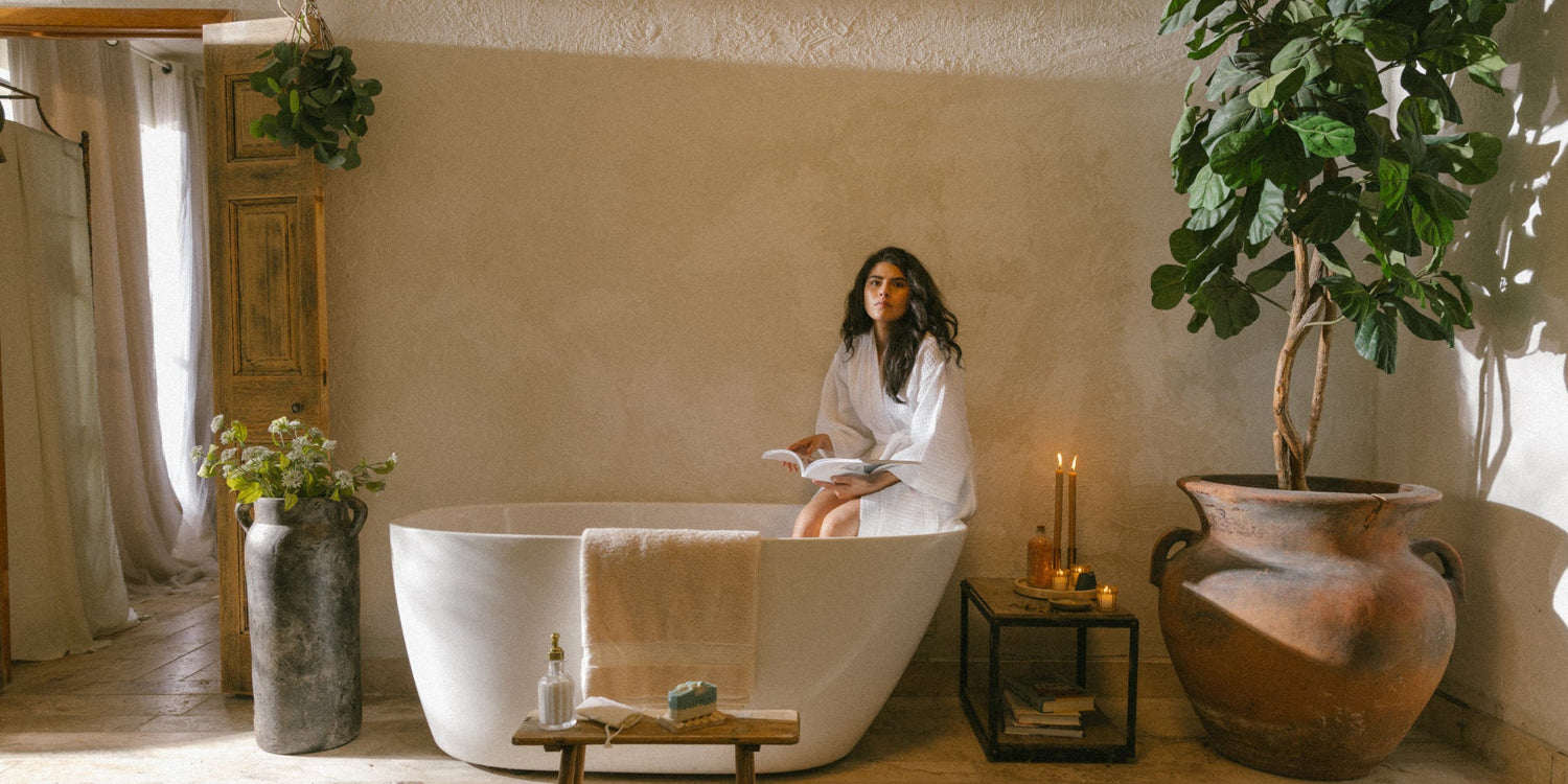 Woman of color relaxing in a luxurious bathtub, reading a magazine, with Oshun goat milk soap displayed on a nearby table for an elegant, clean beauty ritual.