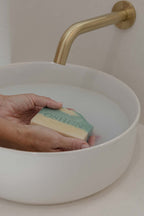 Hand holding a bar of OSHUN soap over a white sink with a gold faucet.