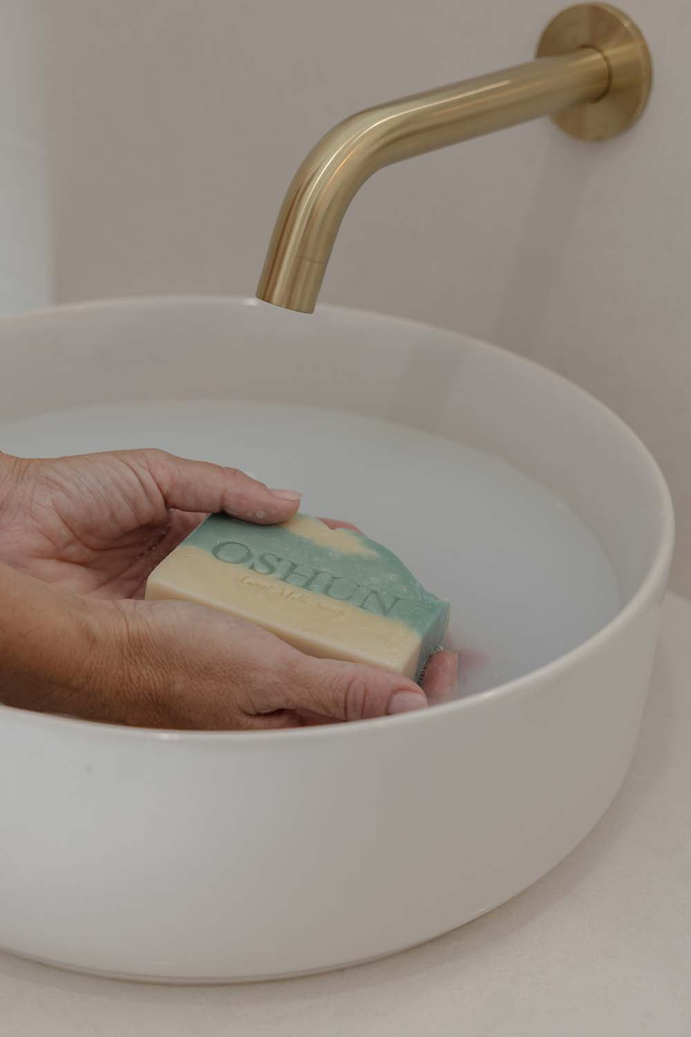 Hand holding a bar of OSHUN soap over a white sink with a gold faucet.