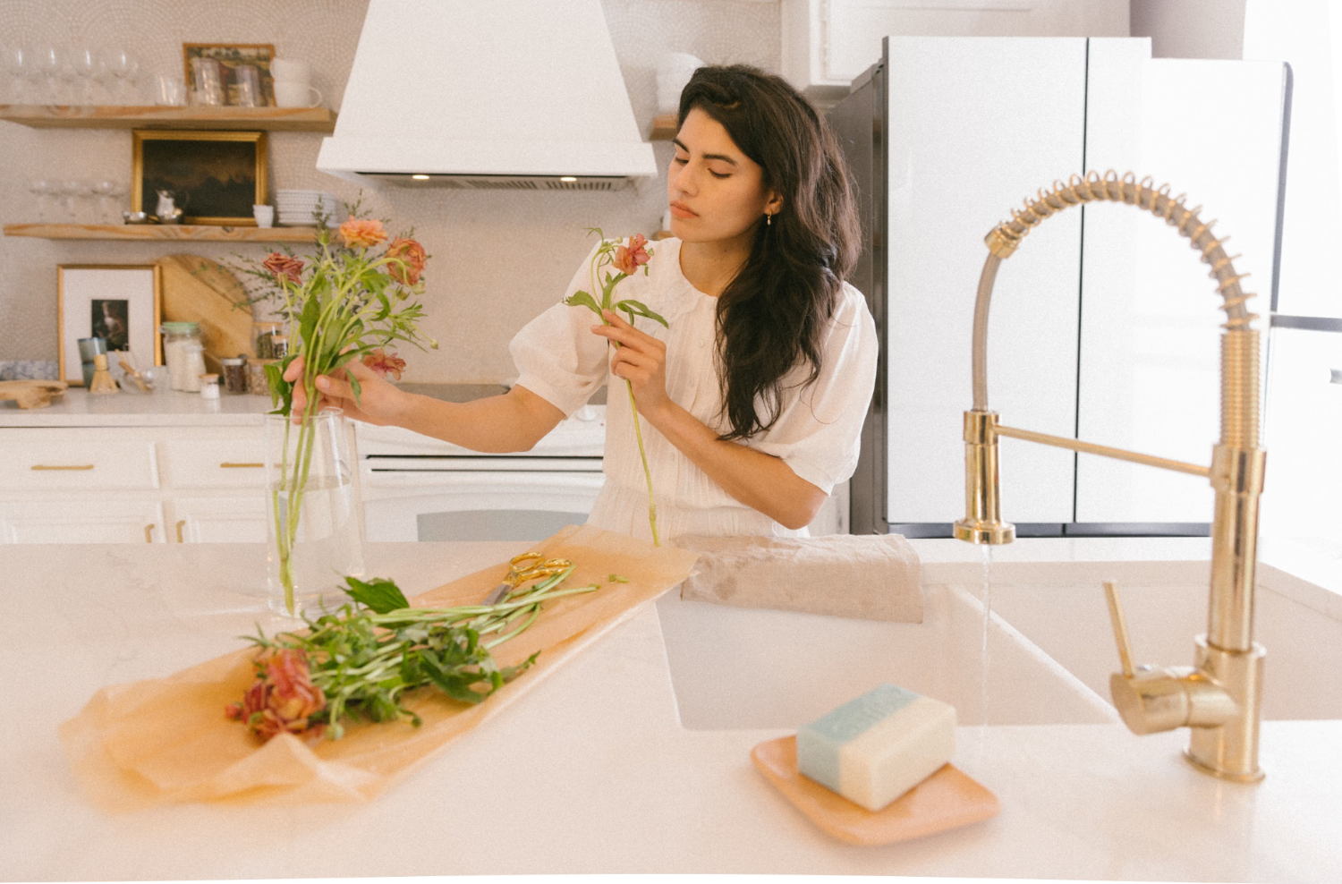 Woman arranging fresh flowers beside Oshun goat milk soap on a kitchen counter—luxury skincare for sensitive skin, fragrance-free, halal beauty.
