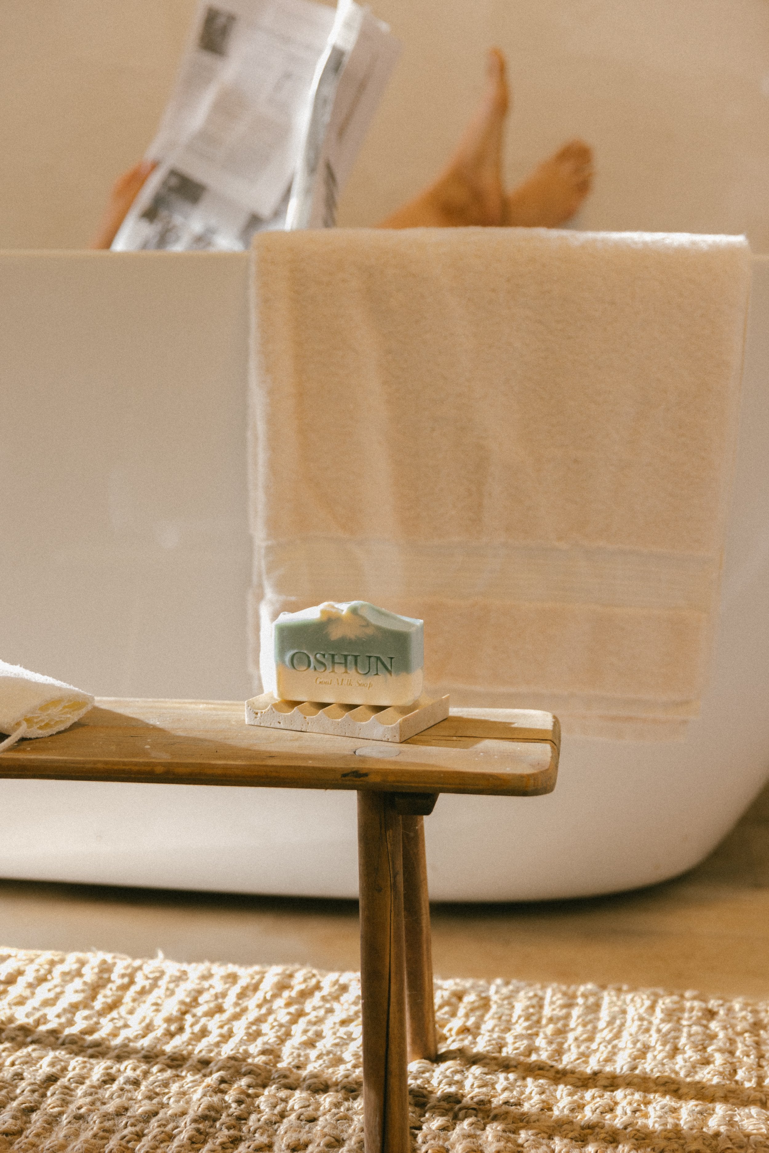 Woman enjoying a spa day with feet up, reading a newspaper, and Oshun goat milk soap resting on a wooden bench—luxury, clean beauty ritual.